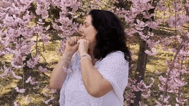 a woman in a white top is standing in front of a tree with pink flowers .