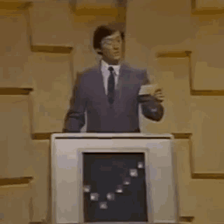 a man in a suit and tie is standing at a podium with a graph on the screen behind him .