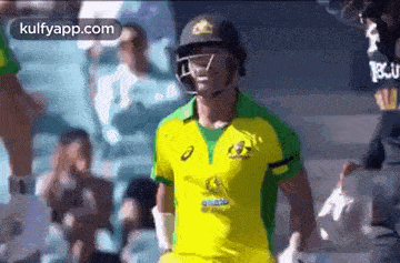a cricket player wearing a helmet and a yellow and green uniform is walking on the field .