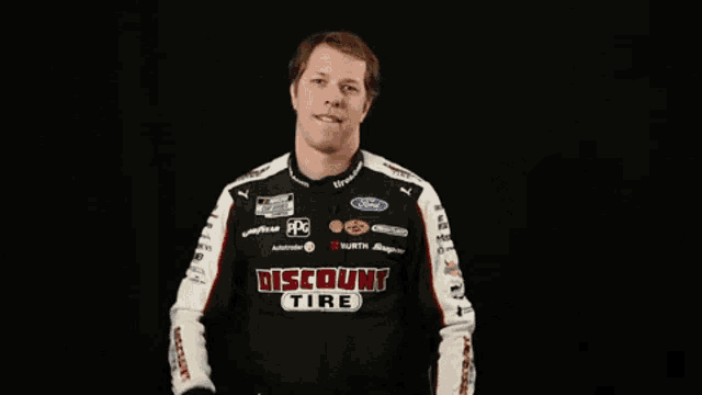 a man in a discount tire racing suit stands in front of a black background