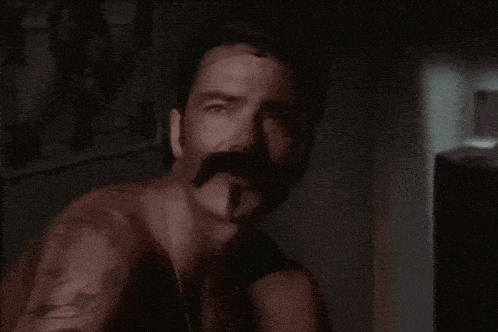 a man with a mustache and a tattoo on his arm is looking at the camera in a dark room .