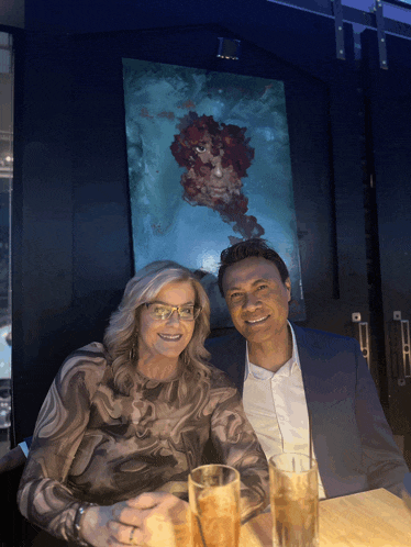 a man and a woman sit at a table with drinks in front of a large painting