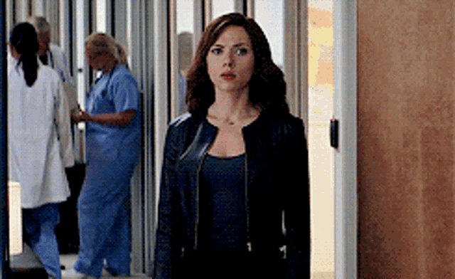 a woman in a black jacket is standing in a hallway with nurses .