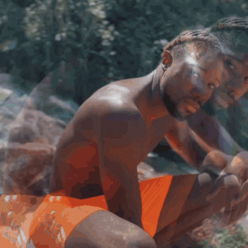 a man without a shirt is sitting in front of a waterfall wearing orange shorts