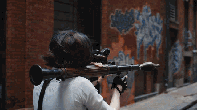 a woman is holding a rocket launcher in front of a brick wall with graffiti