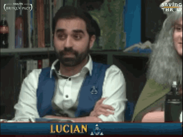 a man with a beard is sitting at a table with his arms crossed in front of a sign that says lucian