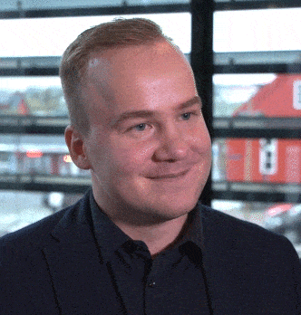 a man in a suit and black shirt smiles in front of a window with the letter e on it