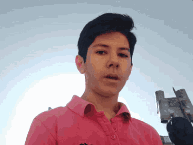a young man wearing a pink polo shirt looks at the camera with a blue sky behind him