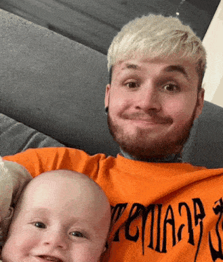 a man with a beard is holding a baby wearing an orange shirt that says ' teman ' on it