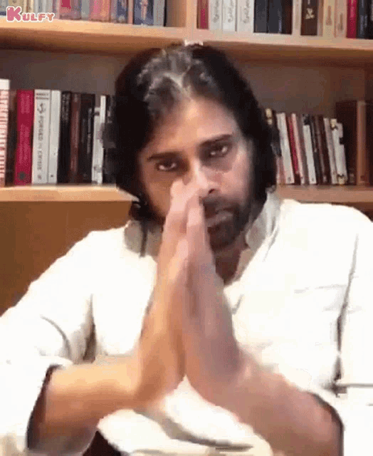 a man is sitting in front of a bookshelf with his hands folded in prayer .
