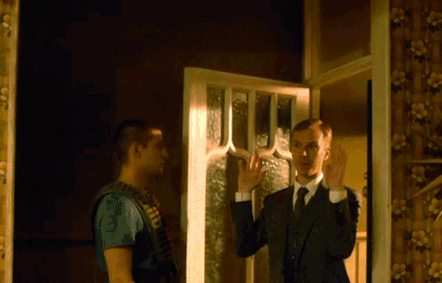 a man in a suit and tie talks to a man in a blue shirt