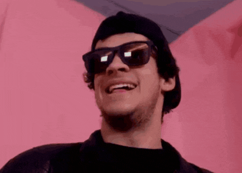 a man wearing sunglasses and a beanie is smiling while standing in front of a pink wall .