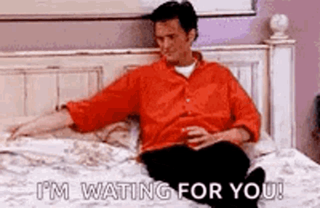 a man in a red shirt is sitting on a bed with a glass of wine .