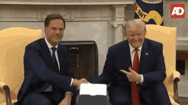 two men in suits and ties are sitting in chairs shaking hands .