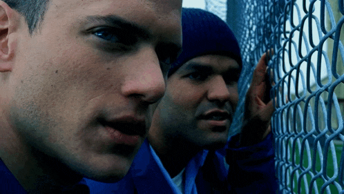 two men are standing behind a chain link fence and one of them is wearing a blue hat