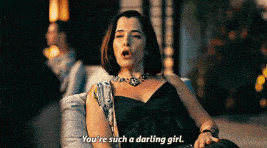 a woman in a black dress and necklace is sitting on a couch and talking to another woman .
