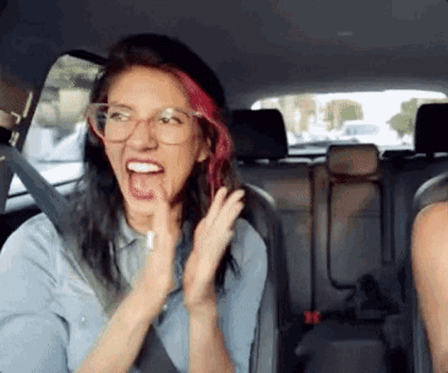 a woman with pink hair and glasses is sitting in the back seat of a car with her mouth open