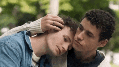 two young men are hugging each other in a park . one of the men is touching the other 's head .