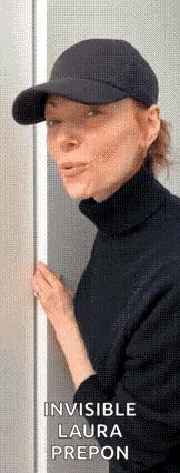 a woman wearing a baseball cap and a turtleneck is standing next to a door and making a funny face .