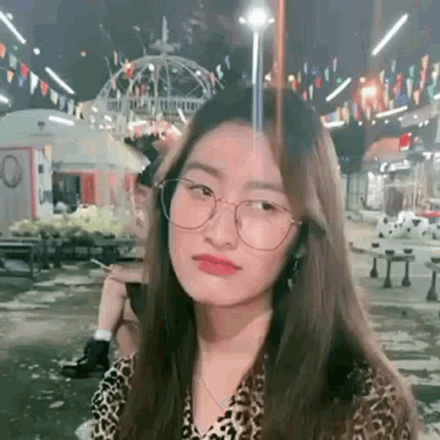 a woman wearing glasses and a leopard print shirt is standing in front of a carnival at night .