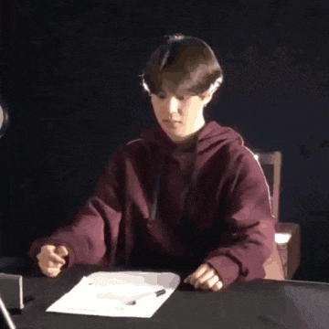 a young man in a maroon hoodie sits at a desk with a piece of paper and a pen .