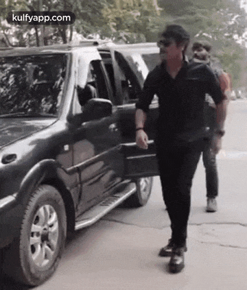 a man in a black shirt is walking towards a black car .