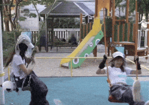 two girls are sitting on a swing in a playground with a slide in the background and one of them is wearing a white hat