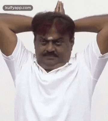 a man with a mustache is wearing a white shirt and making a funny face with his hands behind his head .