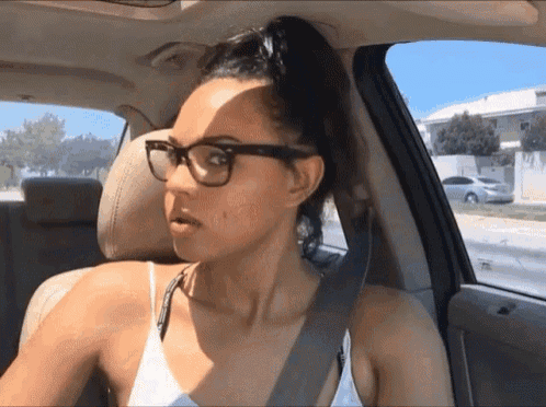 a woman wearing glasses and a white tank top is sitting in the driver 's seat of a car