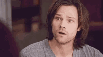 a man with long hair and a beard is making a surprised face while wearing a grey shirt .
