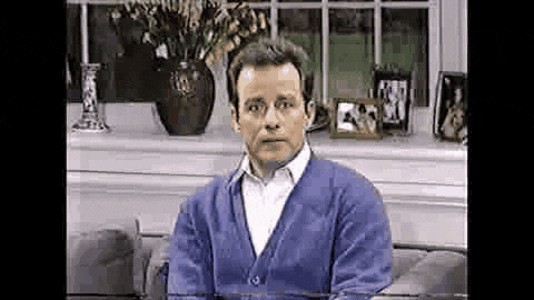 a man in a blue cardigan is sitting on a couch in front of a window .
