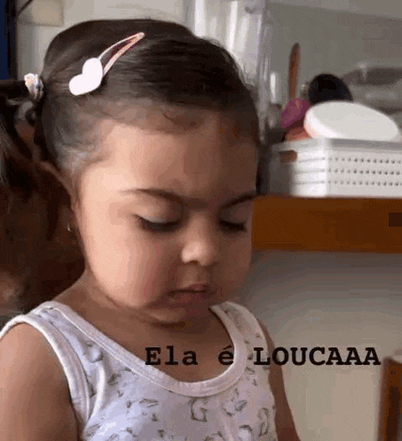 a little girl with a hair clip in her hair is making a funny face while wearing a white tank top .
