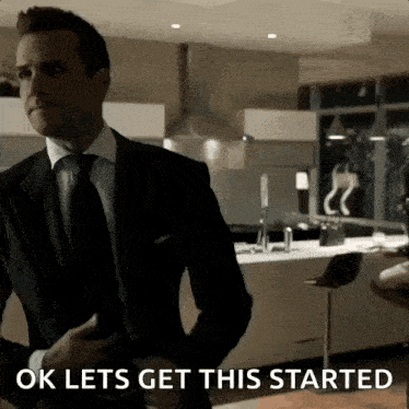 a man in a suit and tie is standing in a kitchen and says ok lets get this started .