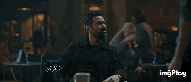 a man with a beard is sitting at a table with a cup of coffee in front of him in a dark room .