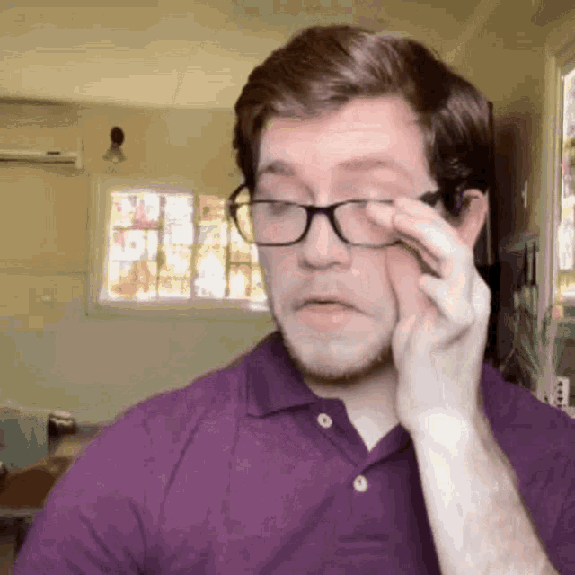 a man wearing glasses and a purple shirt wipes his eye