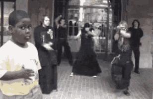 a boy in a yellow shirt stands in front of a group of people dancing on a sidewalk
