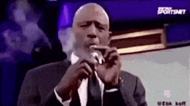 a man in a suit and tie is smoking a cigarette while speaking into a microphone .