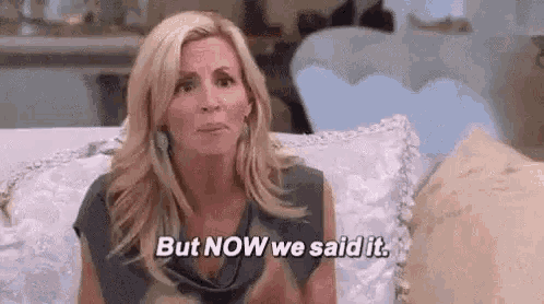 a woman is sitting on a couch with her hands in the air and saying `` but now we said it '' .