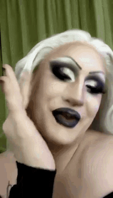 a drag queen with white hair and black lipstick is making a funny face with her hand on her face .