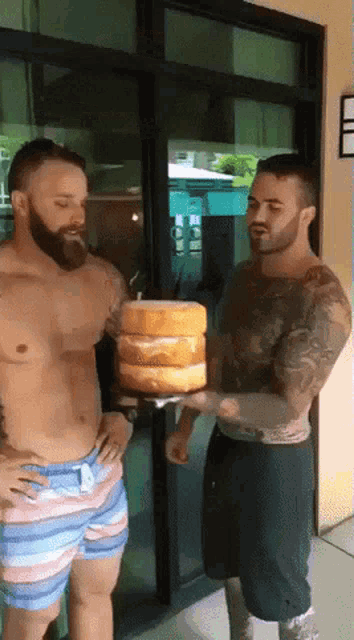 a shirtless man holds a stack of cakes in front of another shirtless man