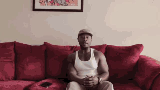 a man in a white tank top sits on a red couch with his hands folded