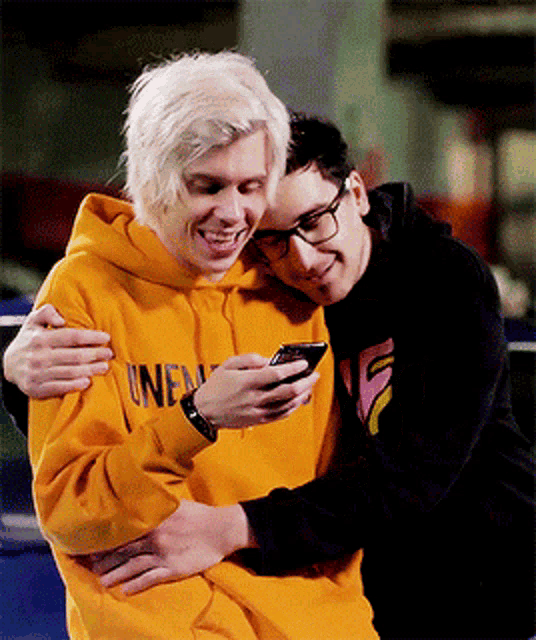 a man in a yellow hoodie is hugging another man while looking at a cell phone