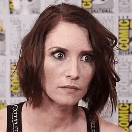 a woman with short brown hair is making a funny face while standing in front of a comic con wall .