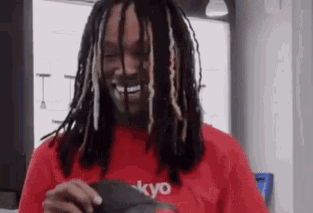 a man with dreadlocks is wearing a red shirt and smiling while holding a cell phone .