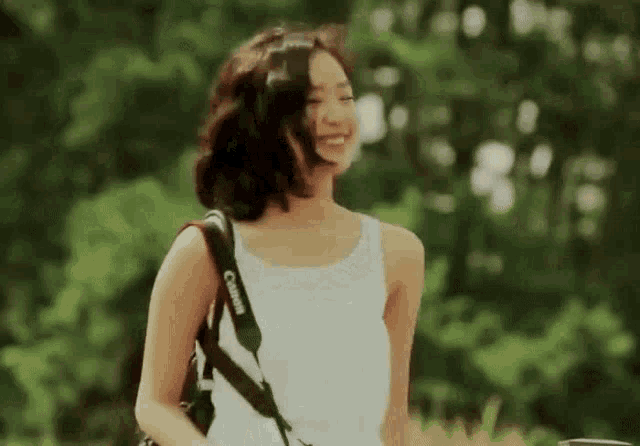 a woman wearing a white tank top and a canon camera strap smiles .