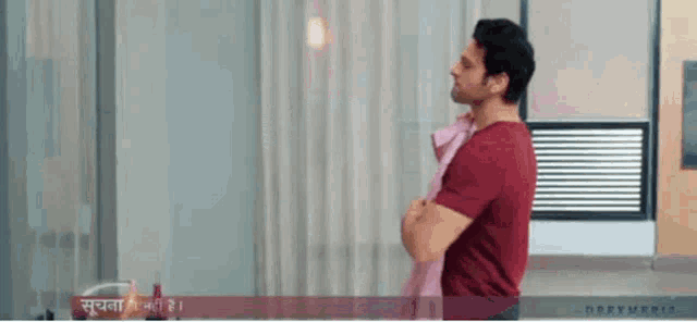 a man in a red shirt is standing in front of a window with his arms crossed and a towel around his neck .