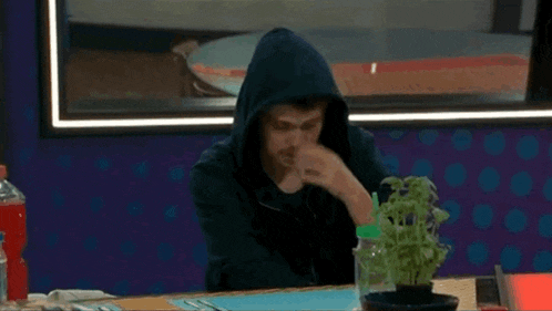 a man in a hooded jacket is sitting at a table with a plant in a pot .