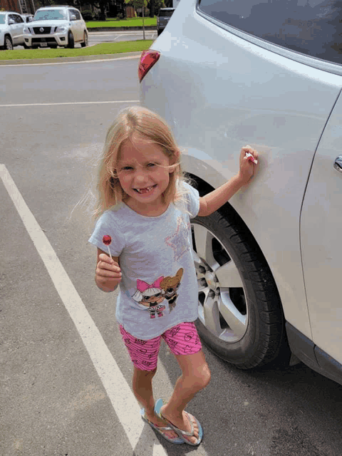 a little girl in a lol surprise shirt is standing next to a car