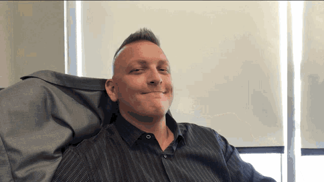 a man in a black shirt is smiling while sitting in a chair in front of a window