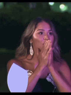a woman is covering her mouth with her hands while wearing a black dress and earrings .